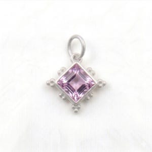May include: A silver pendant with a square, light purple gemstone at its center. The gemstone is surrounded by a silver setting with decorative, rounded edges. The pendant has a small loop at the top for a chain.