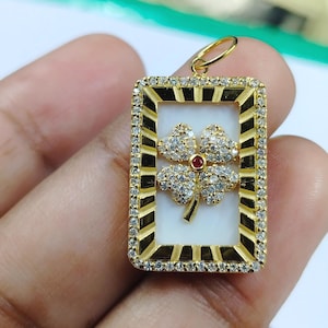Puede incluir: Colgante dorado con un marco rectangular, que encierra un fondo blanco y un diseño de trébol de cuatro hojas. El trébol está adornado con piedras brillantes y una gema roja central. El marco está adornado con pequeñas piedras.