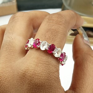 Puede incluir: Un anillo de plata con piedras preciosas ovales alternas en rosa vibrante y blanco transparente. Las piedras están engastadas juntas, creando una banda continua de color. El anillo se muestra en un dedo.