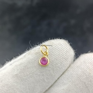 Puede incluir: Un pequeño colgante dorado con una gema rosa redonda. El colgante tiene un diseño sencillo con un lazo para una cadena. La gema está engastada en un bisel dorado. El fondo es de color oscuro y borroso.