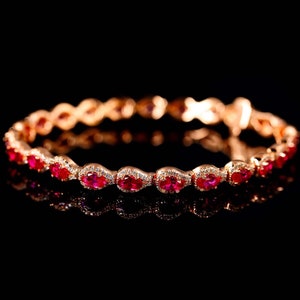 May include: A rose gold bracelet with oval-shaped, vibrant red gemstones and small, clear accent stones. The bracelet is set against a black background, creating a striking contrast that highlights the jewelry's design and sparkle.