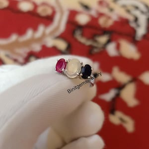 Puede incluir: Un anillo de plata con tres piedras preciosas ovaladas. La piedra central es blanca, la piedra de la izquierda es roja y la piedra de la derecha es azul. El anillo está sobre una superficie blanca.