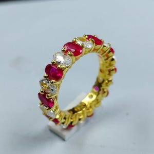 Può includere: Un anello dorato con pietre preziose rosse e chiare alternate. L'anello presenta un design a giro completo, con pietre ovali disposte in modo continuo attorno alla fascia. Le pietre preziose creano un motivo continuo.