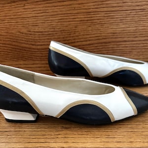 May include: A pair of two-tone flats with a classic design. The shoes feature a white upper with navy blue accents on the toe and heel, and gold trim. The shoes are set on a low heel and are displayed on a wooden surface.