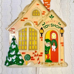 May include: A festive Christmas decoration shaped like Santa's Toy Shop. The building is peach-colored with brick accents, a red door, and a green elf. A green Christmas tree, candy cane, and lollipop are also featured.