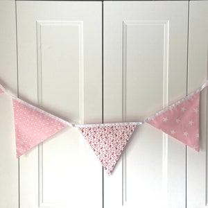 Könnte beinhalten: Eine rosa-weiße Girlande mit verschiedenen Mustern, darunter Punkte, Streifen und Blumenmuster. Die Girlande ist auf einem weißen Band aufgefädelt und hängt an einer weißen Wand.