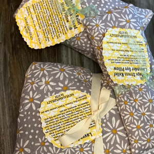 May include: Two gray and white floral print heat packs with yellow daisy designs. The heat packs are tied with a white ribbon and have yellow labels with text that reads "College Stress Relief Lavender Eye Pillow" and "NaturalSimpleCrazy on Etsy".
