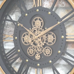 May include: A large, round wall clock with a black face and gold accents. The clock features Roman numerals, gold hands, and visible gears in the center. The outer rim is gold, and the clock has a vintage, industrial design.
