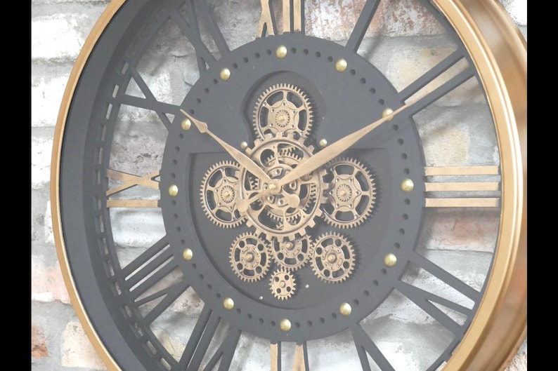 May include: A large, round wall clock with a black face and gold accents. The clock features Roman numerals, gold hands, and visible gears in the center. The outer rim is gold, and the clock has a vintage, industrial design.