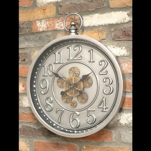 May include: A large, round, silver wall clock with a steampunk design. The clock features large, raised numerals and a decorative centre with gear-shaped elements. The clock is mounted on a brick wall.