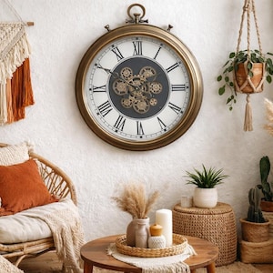 May include: A large, round wall clock with a bronze-coloured frame and Roman numerals. The clock face features visible gears. The room is decorated in a bohemian style, with a woven chair, macrame wall hanging, and potted plants.