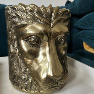 May include: A gold-coloured, lion-faced decorative container. The container is cylindrical with a detailed lion's face sculpted on the front. The lion's mane and facial features are intricately designed, giving it a regal appearance. The container is likely made of metal.