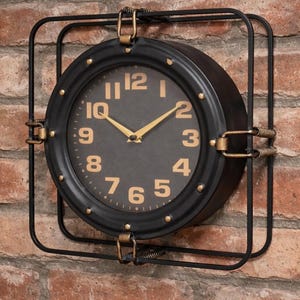 May include: A round wall clock featuring a black circular frame with gold-coloured numerals and hands. The clock is encased within a square black metal wire frame, connected by four gold-toned metal clips at the top, bottom, and sides.