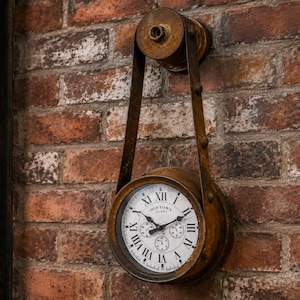 May include: A round wall clock with a weathered bronze finish, suspended by a metal strap from a circular wall mount. The white clock face features black Roman numerals, three small sub-dials, and the text Old Town Clocks in black lettering.