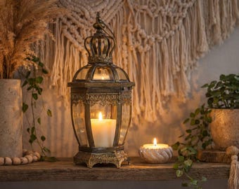 May include: An ornate, antique-style lantern with a lit candle inside. The lantern is gold and glass, with a crown-like top. A smaller candle in a decorative holder and a potted plant are also visible.