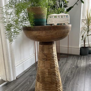 May include: A round wooden side table with a smooth, bowl-shaped top and a tapered, textured base featuring carved horizontal grooves. The table is crafted from natural wood with visible grain patterns and a warm, medium-brown finish throughout.