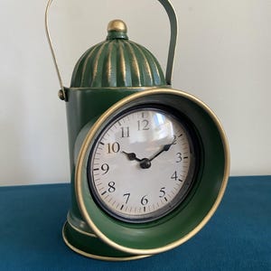 May include: A green metal table clock with a gold handle and a gold trim around the clock face. The clock face has black hands and black Roman numeral markings.