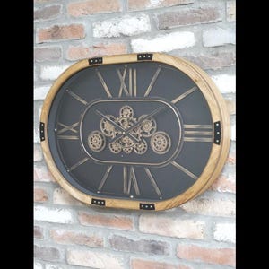 May include: Oval wall clock with a wooden frame and a black face. The clock features gold Roman numerals and a central design of interlocking gears. The clock is mounted on a brick wall.