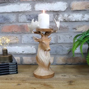 May include: A tan deer head candle holder featuring a sculpted stag bust with light-coloured antlers. A circular platform sits between the antlers, supporting a single white pillar candle that is lit with a visible flame at the top.