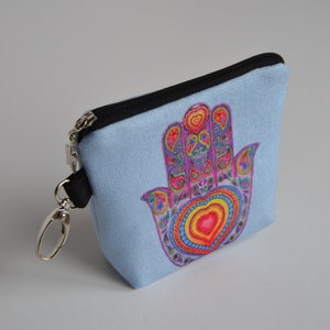 Könnte beinhalten: Hellblaue Reißverschlusstasche mit einem bunten Hamsa-Hand-Design. Die Hamsa-Hand zeigt ein herzförmiges Zentrum und komplizierte Paisley-Muster in Rosa, Gelb, Blau und Lila. Mit silberfarbenem Verschluss und schwarzem Riemen.