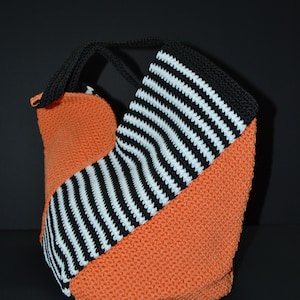 Pode incluir: Bolsa tote de crochê laranja e preta com um padrão de listras diagonais em preto e branco. A bolsa tem uma alça preta e está sobre um fundo preto. Feito à mão.