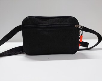 May include: Black fanny pack with a zippered main compartment and a smaller front zippered pocket. The adjustable strap is black, and the zipper pulls are silver. A small orange tag hangs from the zipper.