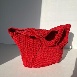 May include: A bright red, crocheted tote bag. The bag has a structured, angular design with integrated handles. The vivid red colour and textured surface create a visually appealing accessory, suitable for various uses.