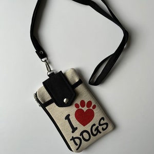 May include: A beige canvas lanyard pouch with a black strap and accents. The pouch features a red heart-shaped paw print and the words "I DOGS" in black. A black flap with a snap closure secures the top.