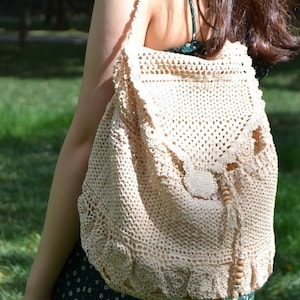 以下が含まれることがあります： クリーム色の手編みバックパック。巾着開閉式で、木製のビーズがアクセント。バッグは、スカラップエッジとフラップクロージャーを備えた、詳細なオープンワークデザインが特徴です。肩にかけて使用します。