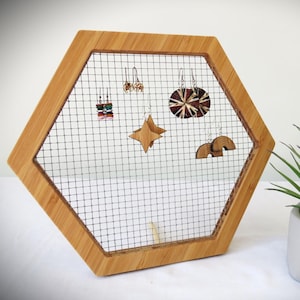 May include: A hexagonal wooden earring holder with a wire mesh insert. The holder has a variety of earrings hanging from it, including a pair of star-shaped earrings, a pair of rainbow-shaped earrings, and a pair of earrings with a circular design.