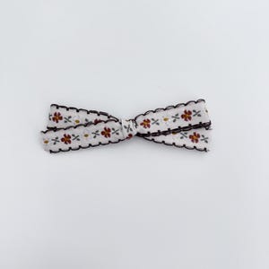 May include: A white ribbon bow with a floral pattern in red, yellow, and brown. The edges are scalloped and outlined in black. This decorative bow is suitable for various craft projects and gift wrapping.