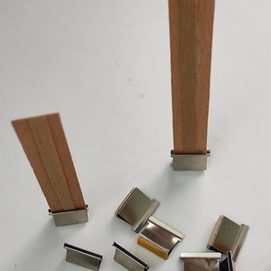 May include: Two wooden candle wicks with metal holders, and a pile of metal candle wick holders. The holders are silver and have a slight shine.