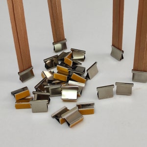 May include: A close-up of a pile of silver metal clips with adhesive backing. The clips are designed to hold fabric together. There are two wooden dowels in the background.