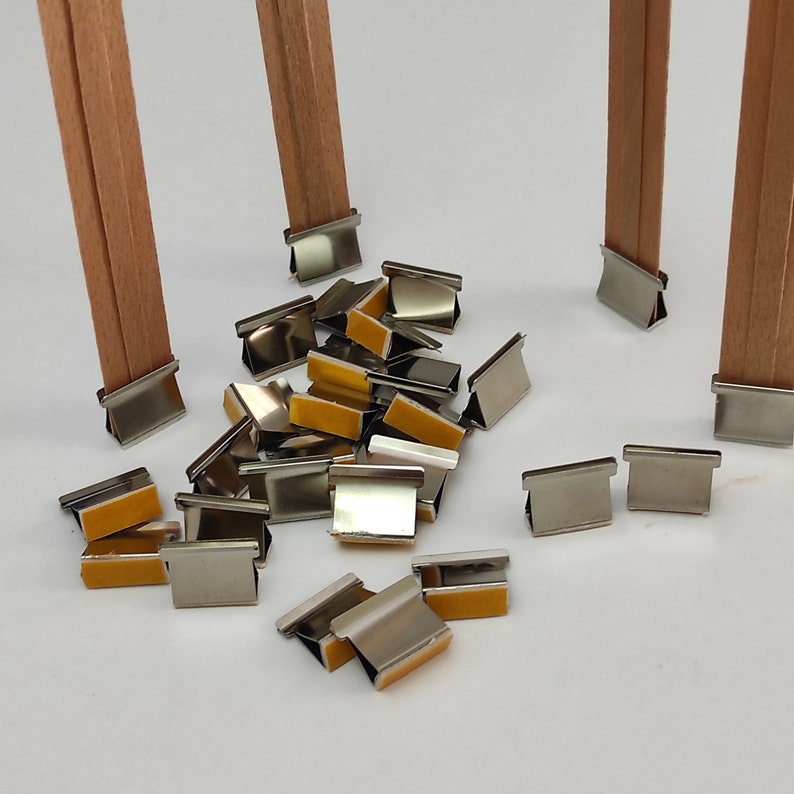 May include: A close-up of a pile of silver metal clips with adhesive backing. The clips are designed to hold fabric together. There are two wooden dowels in the background.