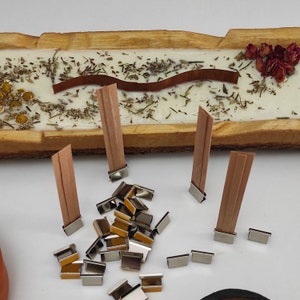 May include: A close-up of wooden candle wicks with metal wick stickers. The wicks are arranged on a wooden surface with dried herbs and flowers.