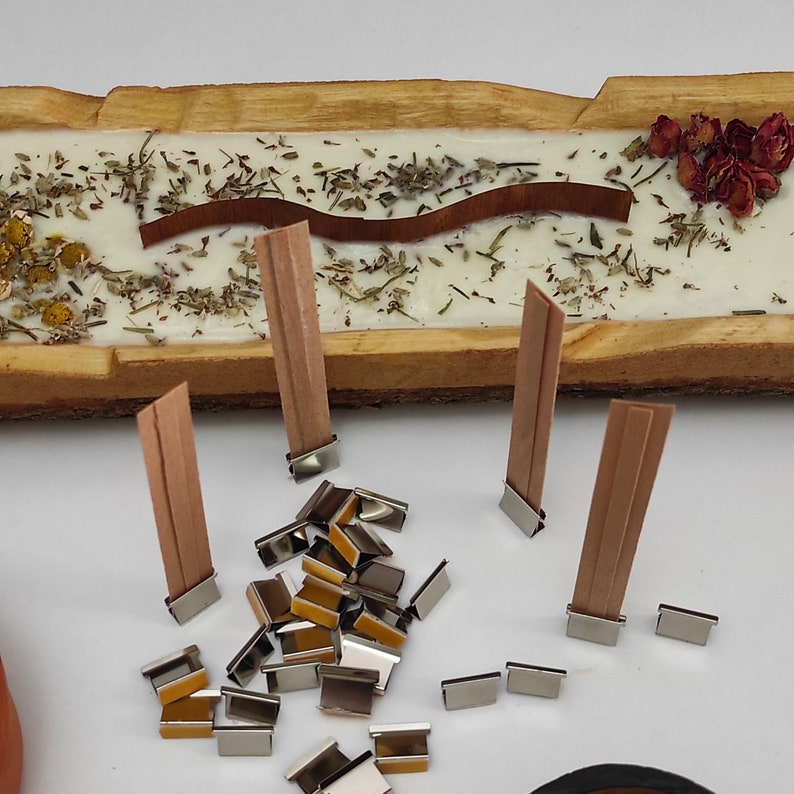 May include: A close-up of wooden candle wicks with metal wick stickers. The wicks are arranged on a wooden surface with dried herbs and flowers.