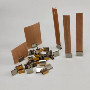 May include: A set of silver metal binder clips with a brown paper backing. The clips are scattered on a white surface. There are also three brown paper strips standing upright in the background.