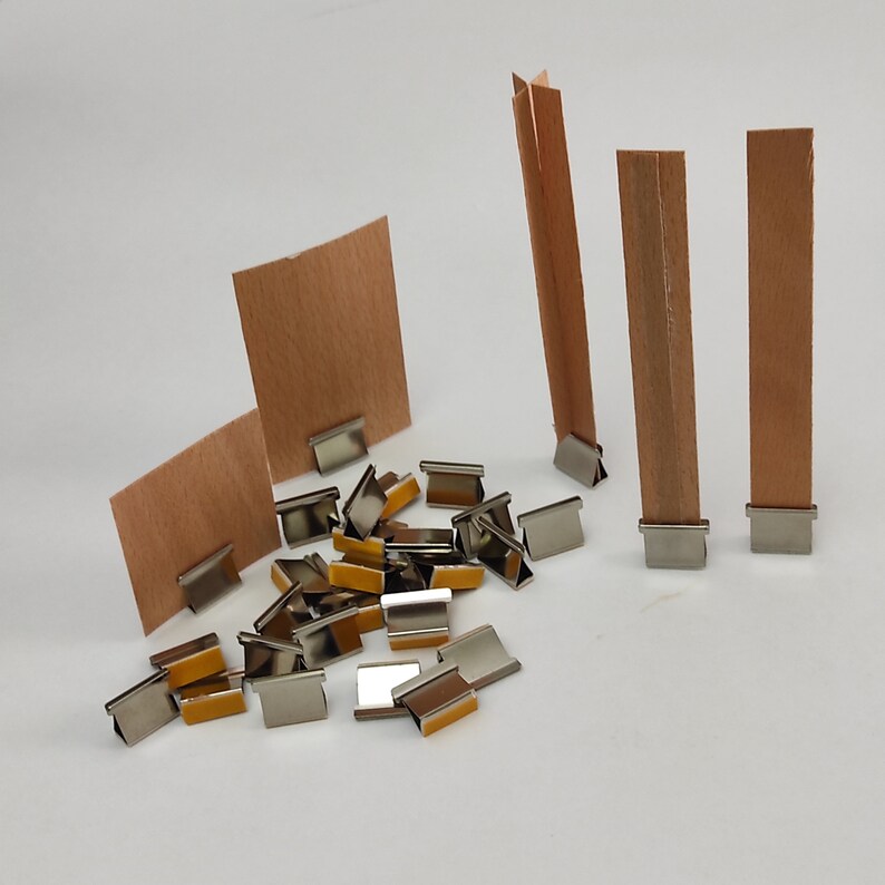 May include: A set of silver metal binder clips with a brown paper backing. The clips are scattered on a white surface. There are also three brown paper strips standing upright in the background.