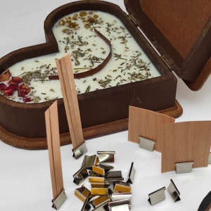 May include: A wooden heart-shaped box with a lid open, revealing a white candle with dried herbs and flowers inside. The box is surrounded by wooden wicks and metal candle clips.