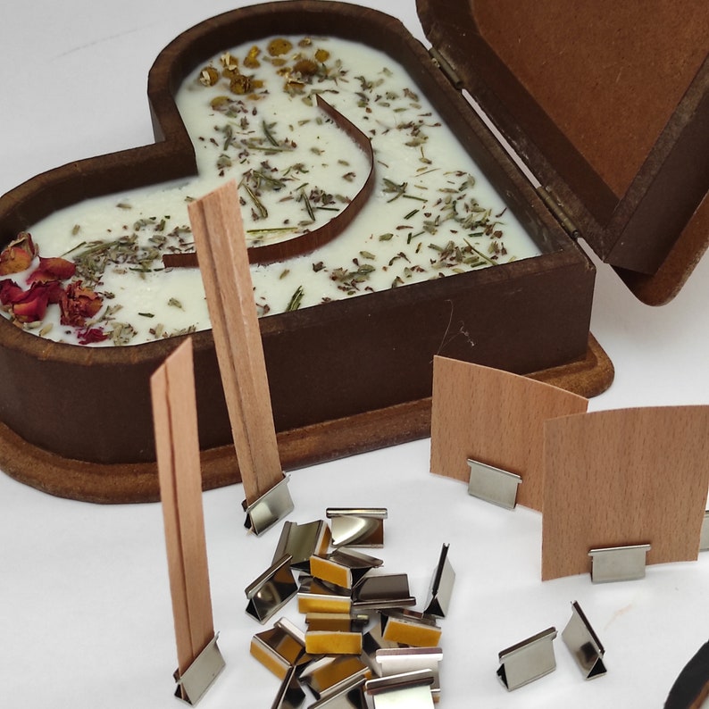 May include: A wooden heart-shaped box with a lid open, revealing a white candle with dried herbs and flowers inside. The box is surrounded by wooden wicks and metal candle clips.