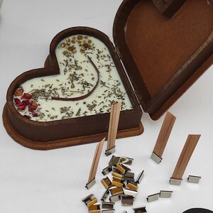 May include: A heart-shaped wooden box with a lid, containing a white candle with dried herbs and flowers. The box is open and there are several wooden wicks and metal candle clips on a white surface.