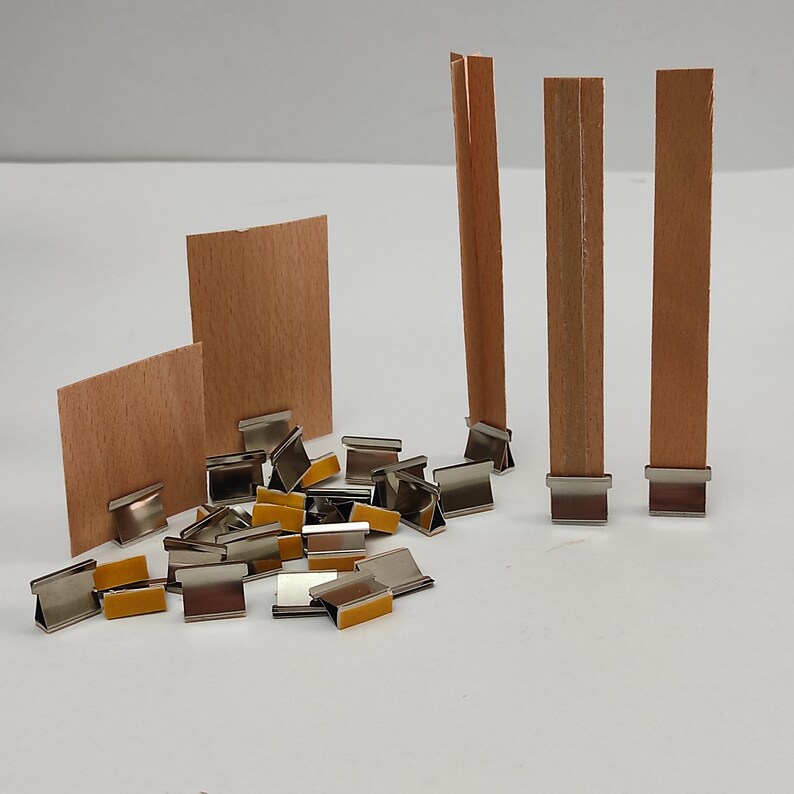 May include: A set of silver metal clips with adhesive pads on the bottom. The clips are used to hold wooden candle wicks in place. There are three wooden wicks standing upright in the background.