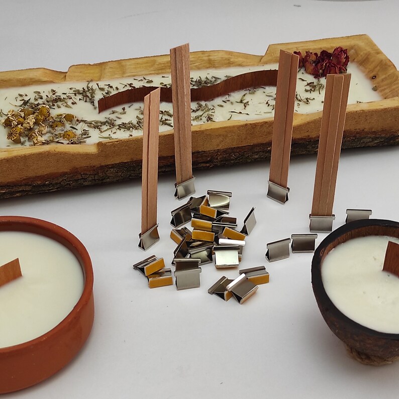 May include: A close-up of wooden candle wicks and metal wick stickers. The wicks are brown and the stickers are silver with a yellow adhesive.