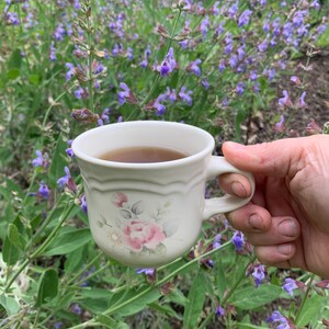 May include: A white ceramic mug with a floral design, filled with tea, held in front of a field of purple sage flowers.