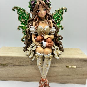 May include: A fairy figurine with long brown hair, a green leaf crown, and large green wings. The figure wears a cream-coloured dress and holds a small orange fox. The fairy has white stockings with gold moon and star patterns and copper-coloured shoes.