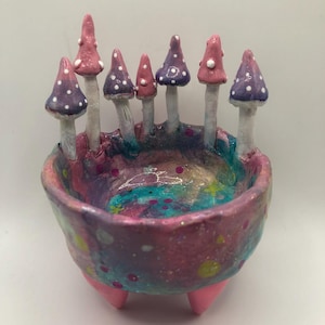May include: A colourful ceramic bowl with a galaxy-themed interior and exterior. The bowl is decorated with several mushroom figures with pink and purple caps, each with white polka dots. The bowl has three pink feet.