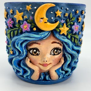 May include: A cylindrical ceramic planter featuring a hand-painted relief of a face with flowing blue hair, resting hands, and a crescent moon crown. The dark blue background is decorated with yellow stars, green vines, and small purple flowers.