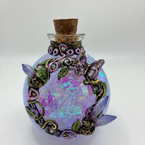 May include: A decorative glass bottle with a cork stopper. The bottle is filled with iridescent material and adorned with a butterfly, leaves, and crystals. The bottle has a purple and pink colour scheme with gold accents.