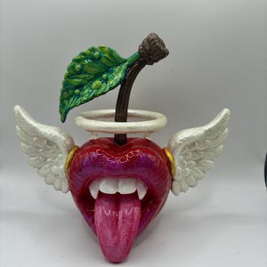 May include: A sculpture of a red cherry with a human-like mouth featuring white teeth and a protruding pink tongue. The cherry has a green leaf, a brown stem, and two white feathered wings extending from the sides with a white halo above.
