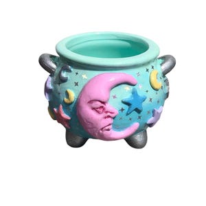 May include: A teal cauldron with silver handles and feet. The cauldron is decorated with a pink crescent moon with a face, blue stars, and yellow and purple crescent moons. The interior of the cauldron is teal.